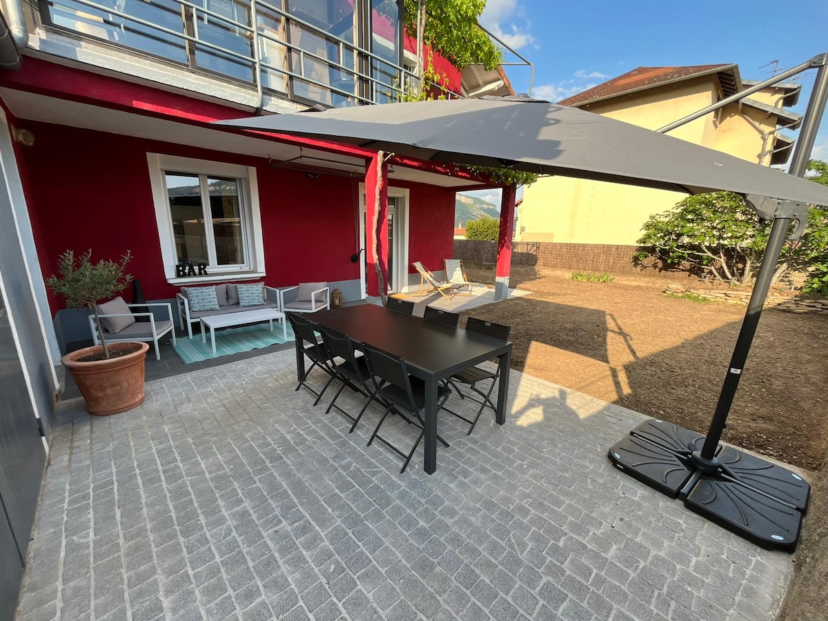 An outdoor patio area features a large black dining table surrounded by chairs. A shaded lounge area with two reclining chairs is situated nearby. The wall is painted red, with a window offering a view of the garden space, complemented by potted plants.