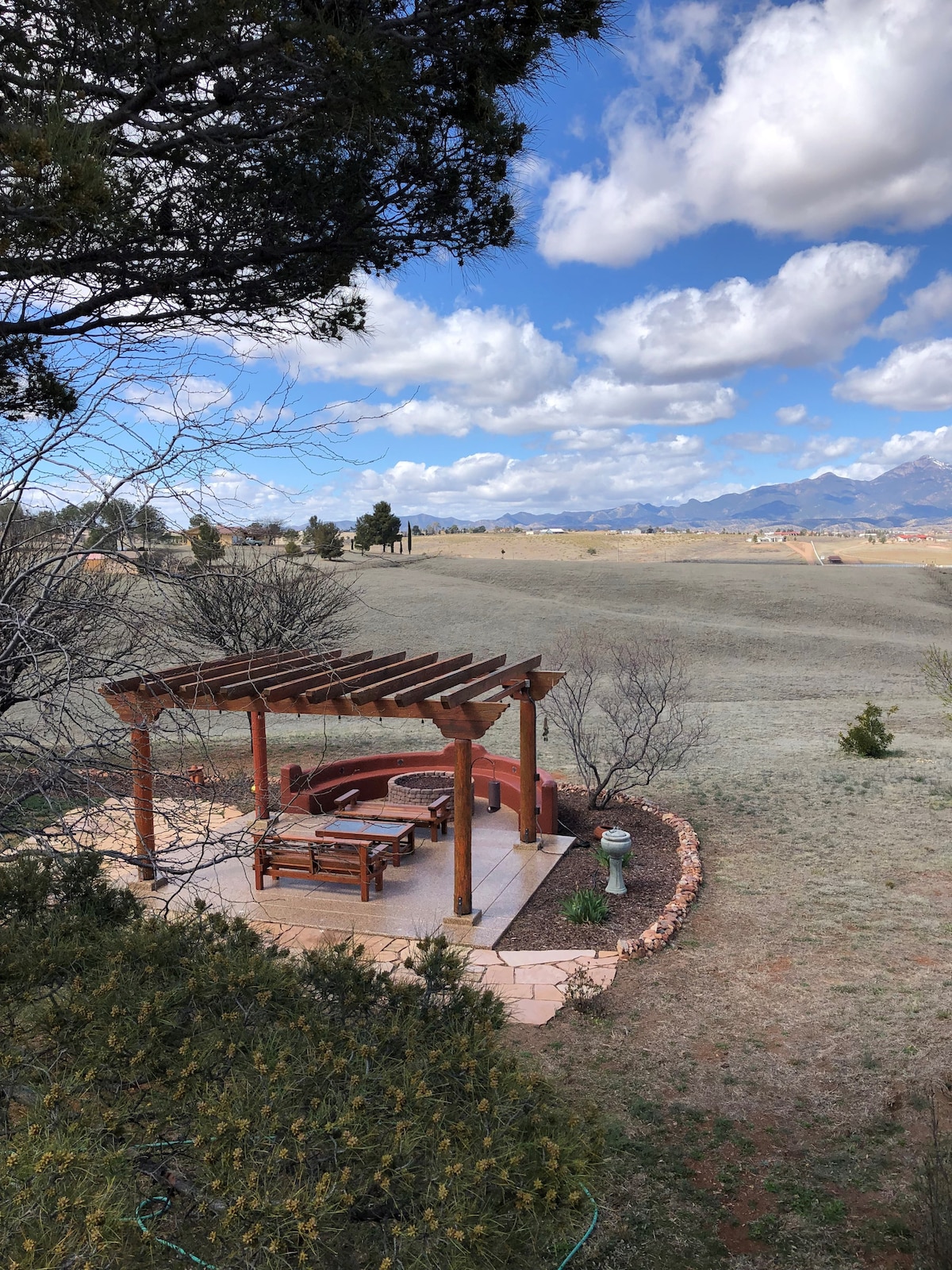 An outdoor patio area is highlighted by a wooden pergola, offering shaded seating around a circular stone fire pit. Open grasslands extend in the background, with distant mountains visible under a partly cloudy sky.