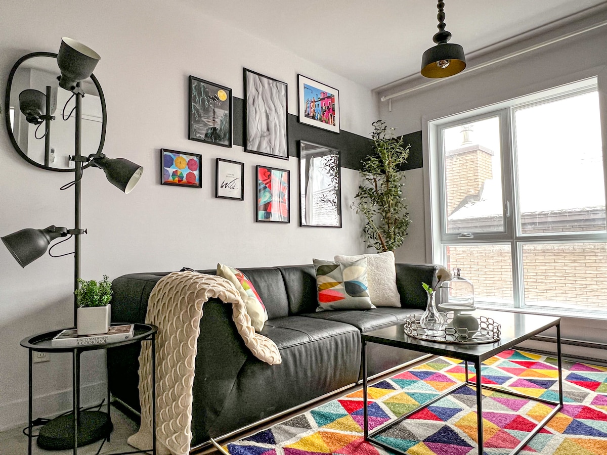 A modern living area features a spacious black sofa adorned with colorful cushions. A stylish coffee table sits on a vibrant geometric rug. Wall art enhances the atmosphere, and natural light streams through a large window, complemented by a potted plant and contemporary floor lamp.