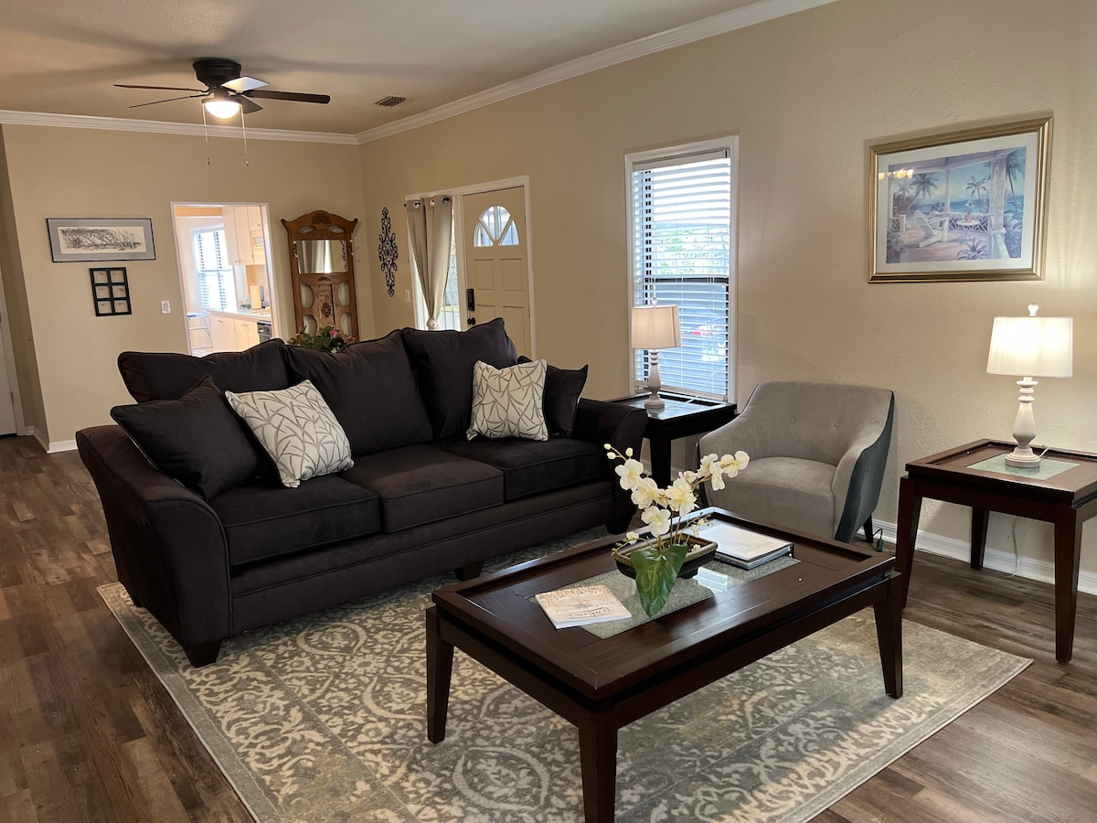A cozy living area features a dark upholstered sofa adorned with patterned throw pillows and a light accent chair. A coffee table displays a decorative arrangement, while soft lamps provide warm lighting. Natural light enters through the windows, enhancing the inviting space.