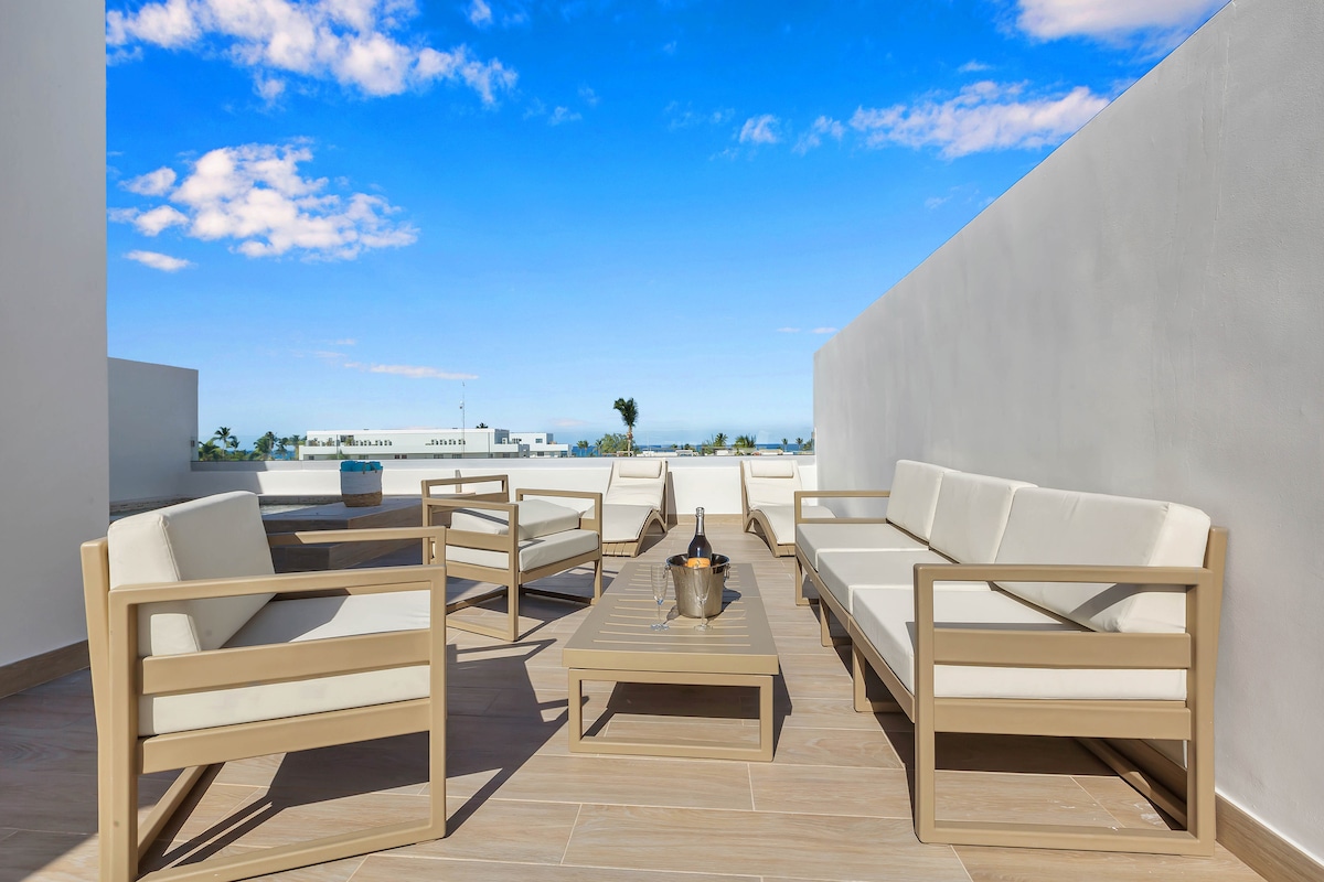 A spacious terrace is furnished with comfortable white lounge chairs and a low table, creating an inviting outdoor seating area. The bright blue sky and surrounding greenery can be seen in the background, enhancing the relaxed atmosphere of the penthouse.