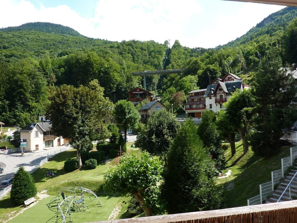 The image shows a scenic view of a lush green landscape, featuring a playground in the foreground and residential buildings in the background. A chairlift ascends the mountainside, while a rich canopy of trees occupies the hillside, creating a serene, inviting environment.