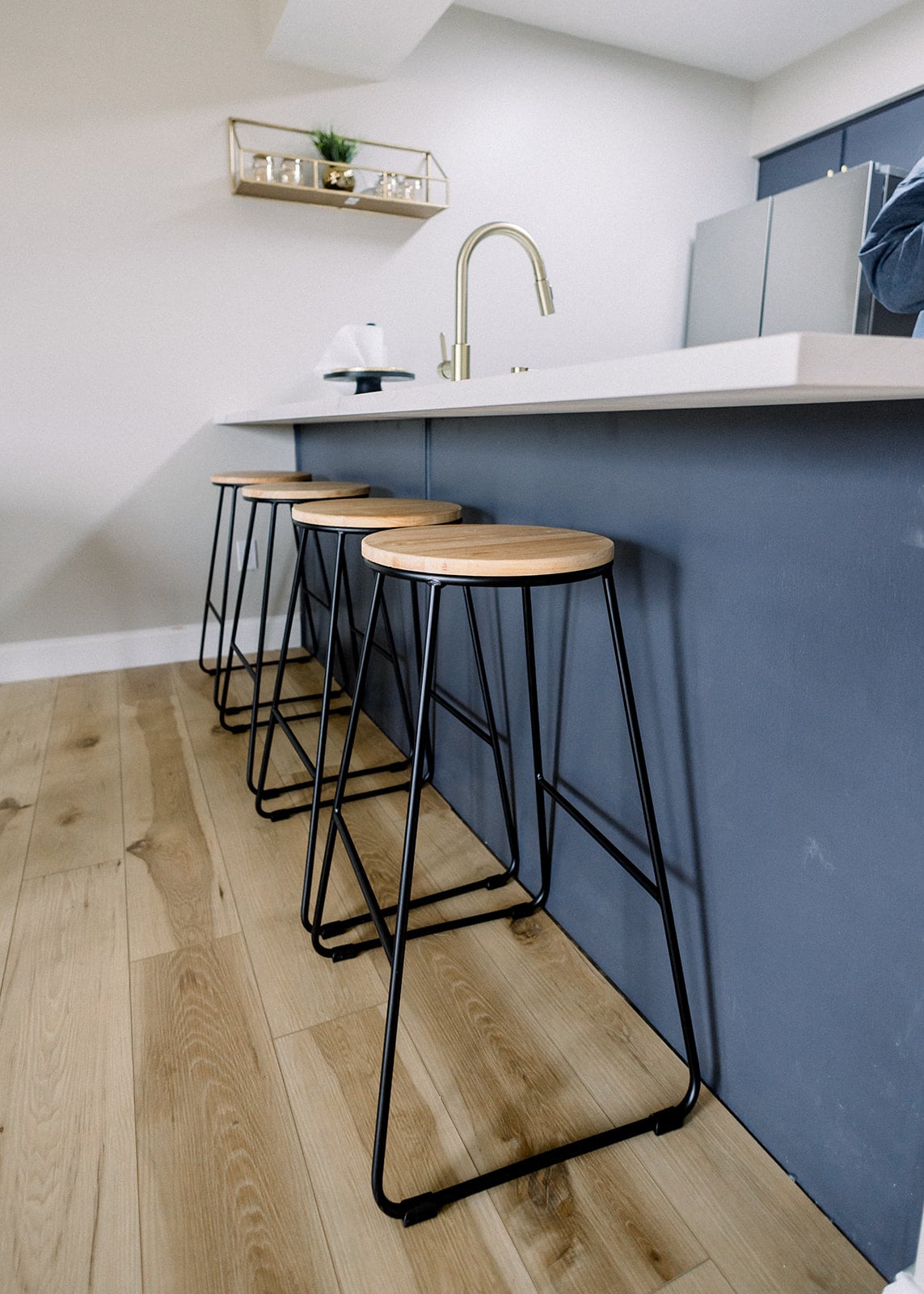Three sleek bar stools are positioned along a modern kitchen island, featuring wooden seats supported by black metal frames. The light wood flooring adds a warm touch to the contemporary space, while a minimalist shelf with greenery is visible in the background.