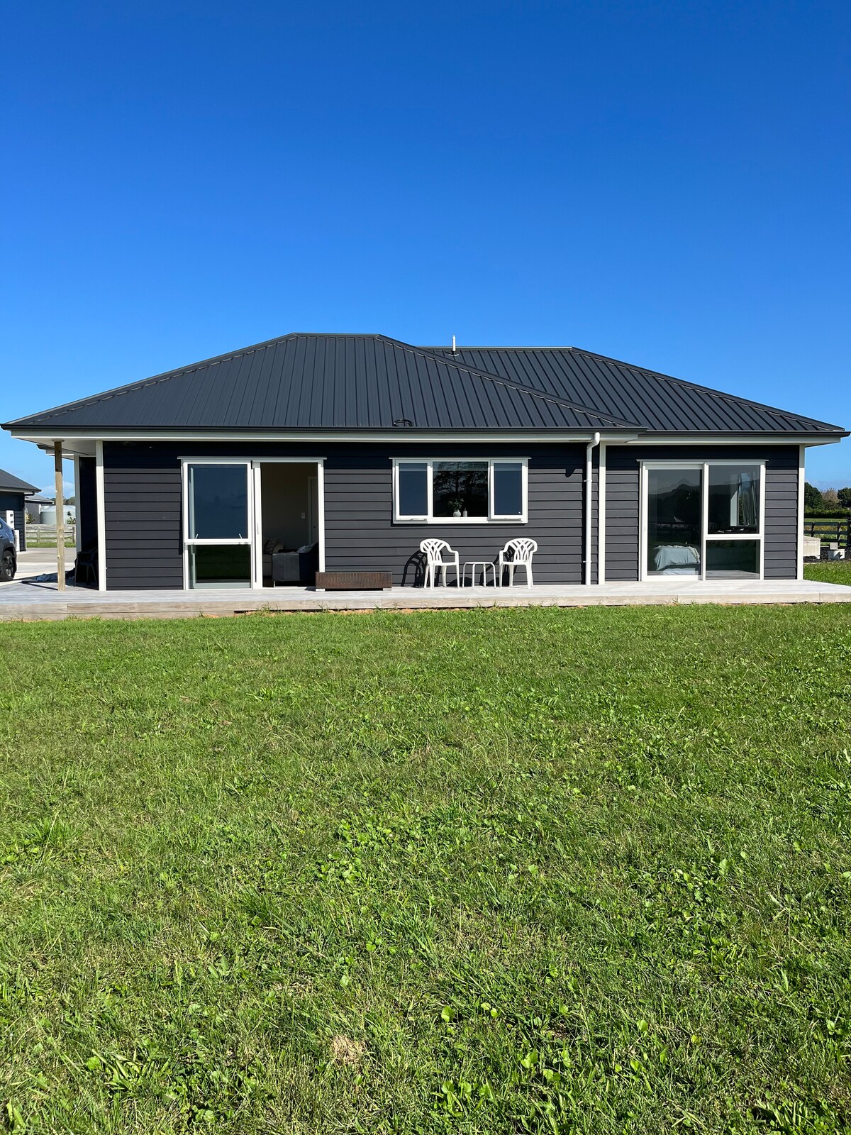 The exterior of a modern one-story unit is visible, featuring a dark grey facade and a pitched roof. Two chairs are positioned on a small patio, with large windows allowing natural light to enter the interior. A vast green lawn surrounds the property.
