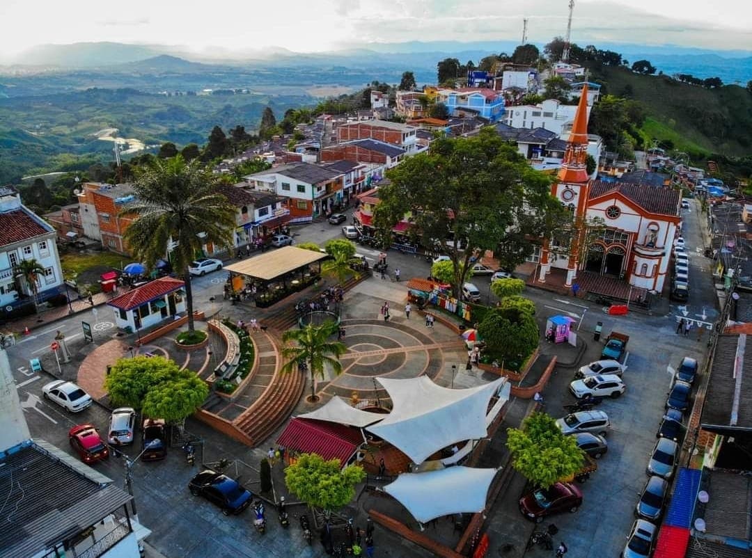 An aerial view captures a vibrant town square surrounded by trees and parked vehicles. The square features a circular design, with diverse structures, including a church with an orange spire. Rolling hills and a scenic horizon are visible in the background.