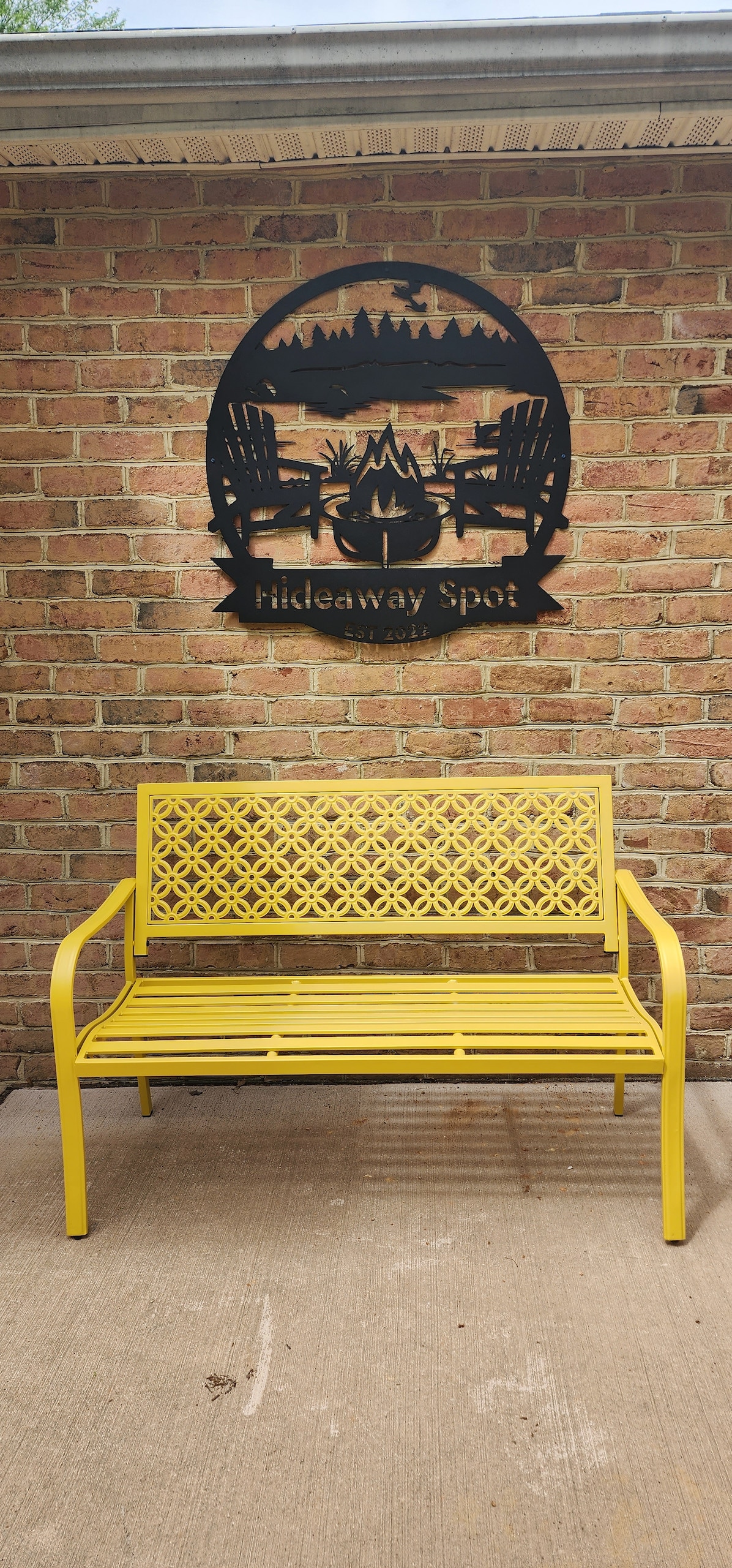 A vibrant yellow bench is positioned against a textured brick wall, featuring a decorative metal sign above that reads 'Hideaway Spot.' The bench's intricate design adds a welcoming touch to the outdoor space.
