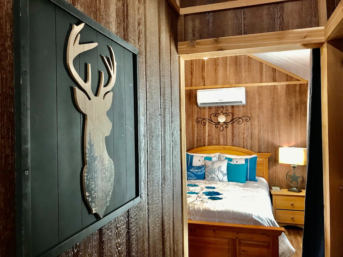 A wooden plaque featuring a deer silhouette hangs on a wall, leading toward a bedroom furnished with a wooden bed. The bed is adorned with blue and white decorative pillows, while a lamp provides warm lighting beside a wooden nightstand.