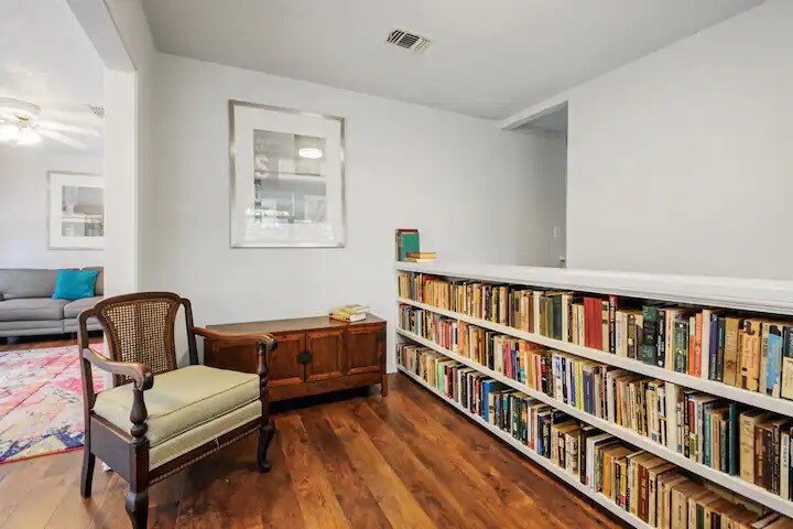 A cozy reading nook features a honey-colored armchair beside a wooden side table. A bookshelf filled with assorted books runs along the wall, complemented by light wood flooring. Natural light enhances the neutral tones of the space, fostering a calm and relaxing environment.
