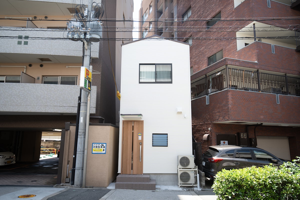 A two-story private house is presented in a modern architectural style. The exterior features a clean, white facade, large windows, and a wooden door. Surrounding elements include parked vehicles and nearby buildings, contributing to the urban setting.
