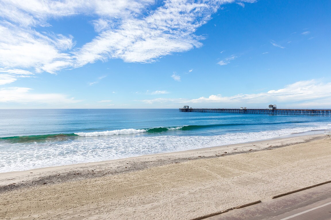 A serene beach view features gentle waves lapping against the shore, with soft sand stretching along the coastline. In the distance, the Oceanside Pier extends into the water under a clear blue sky, creating a tranquil seaside environment.