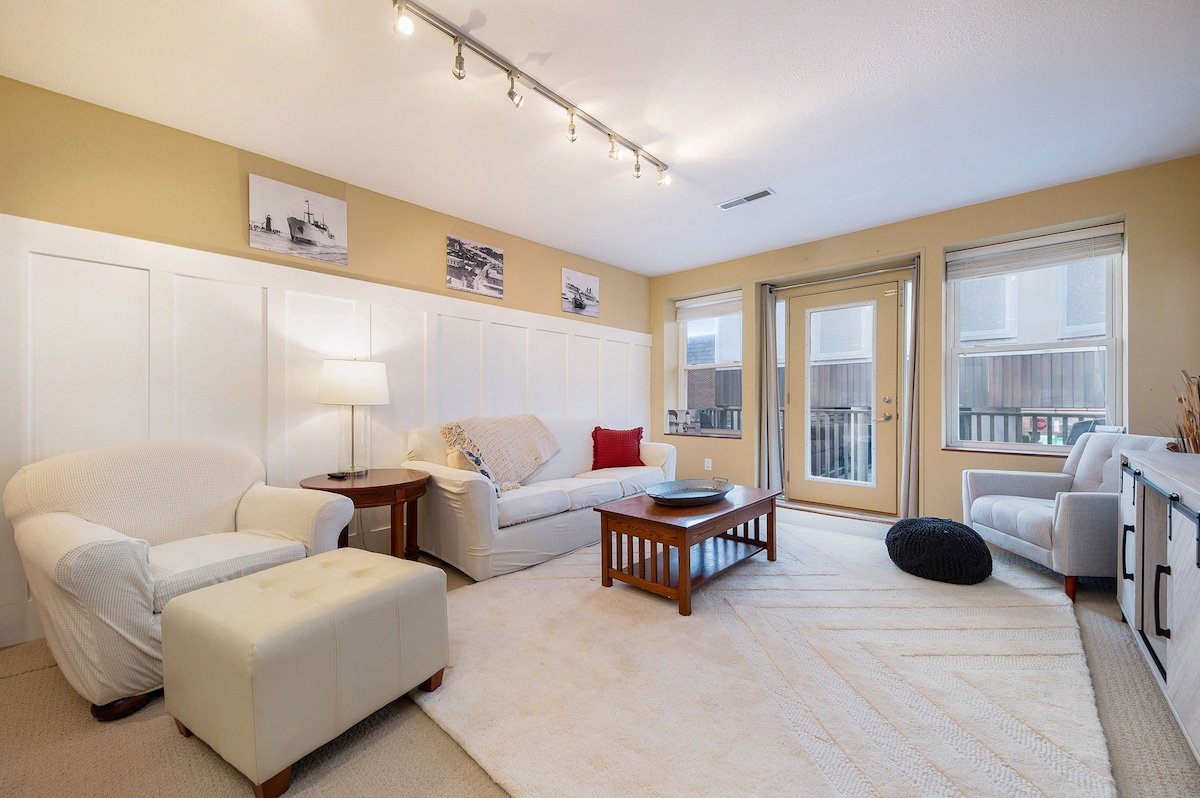 A bright living area features a light-colored sofa and armchairs arranged around a wooden coffee table. Soft lighting is provided by track lights above, and large sliding glass doors lead to a balcony. A textured area rug adds warmth to the space.