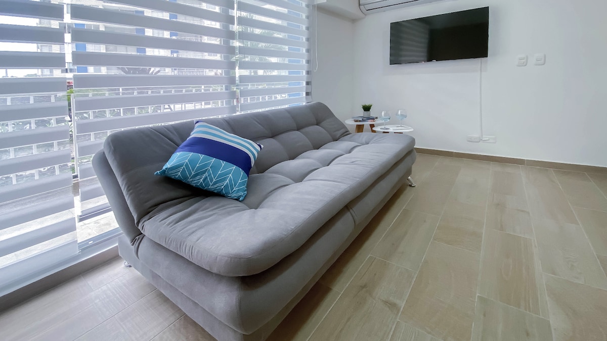 A comfortable sofa bed is positioned in a well-lit living area, complemented by light-colored flooring. A flat-screen TV is mounted on the wall, while sheer blinds allow natural light to filter in softly. A small side table adds a functional touch.