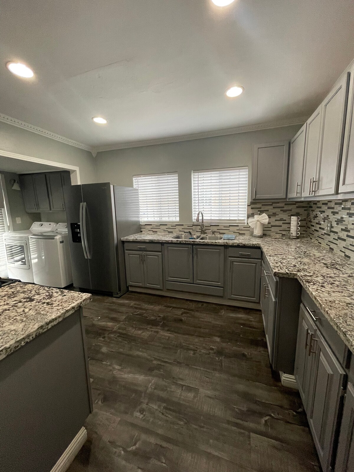 The kitchen features modern cabinetry and a spacious layout. Grey granite countertops contrast with light grey cabinets, while a large stainless steel refrigerator stands beside a sink under two windows. Natural light illuminates the room, complemented by recessed lighting on the ceiling.
