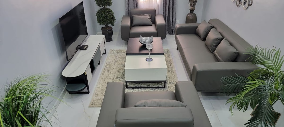 A stylish living room is presented with modern furniture in neutral tones. Two sofas and a lounge chair are arranged around a central coffee table. Lush plants and decorative accents add a touch of greenery, while a television stands against one wall under a large window.
