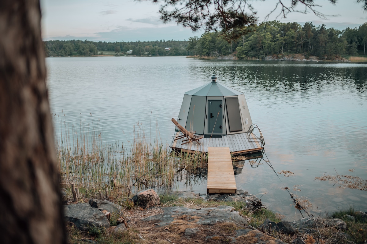 A circular hut is perched on a floating platform over calm waters, surrounded by lush greenery and natural landscapes. A wooden walkway leads to the entrance, while a sun lounger rests nearby, providing a serene spot to enjoy the panoramic views of the surrounding area.