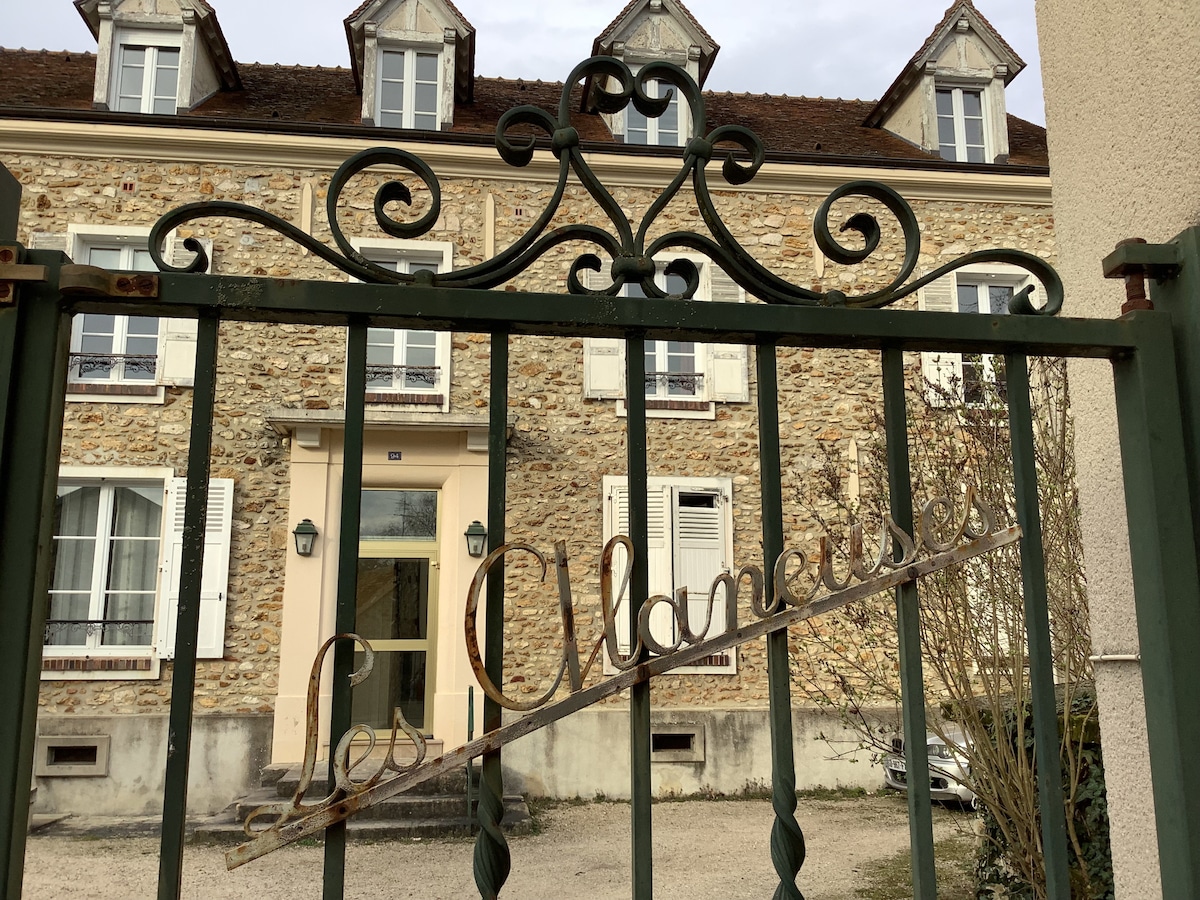 An ornate iron gate, adorned with the name 'Les Châtaigniers,' leads to a charming stone building. The facade features multiple windows with white shutters and a welcoming entrance, bordered by a gravel pathway and neatly trimmed shrubs.