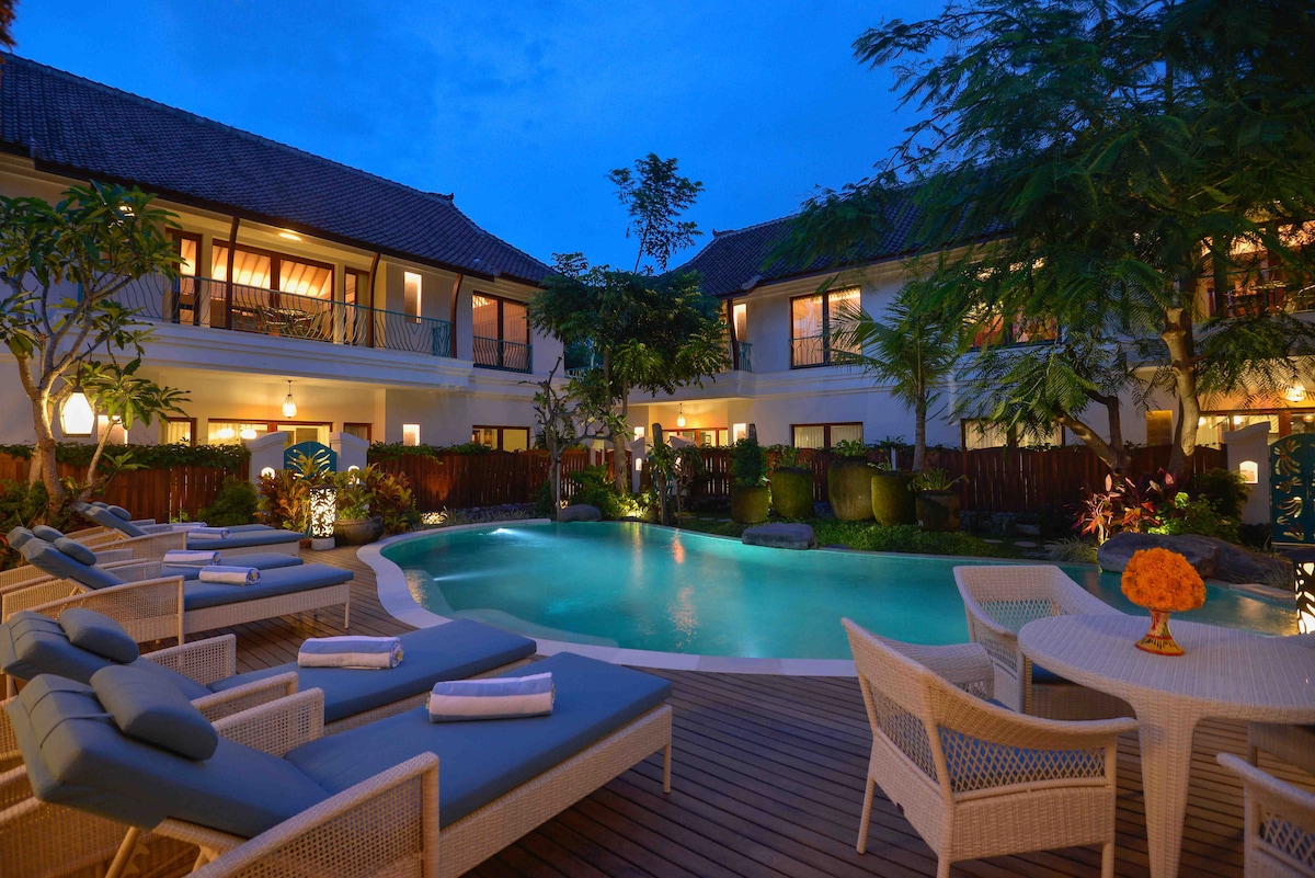 The outdoor space features a serene swimming pool surrounded by sun loungers and tables. Lush greenery and thoughtfully placed lighting enhance the tranquil ambiance, as the building's architecture provides a cozy backdrop under the evening sky.
