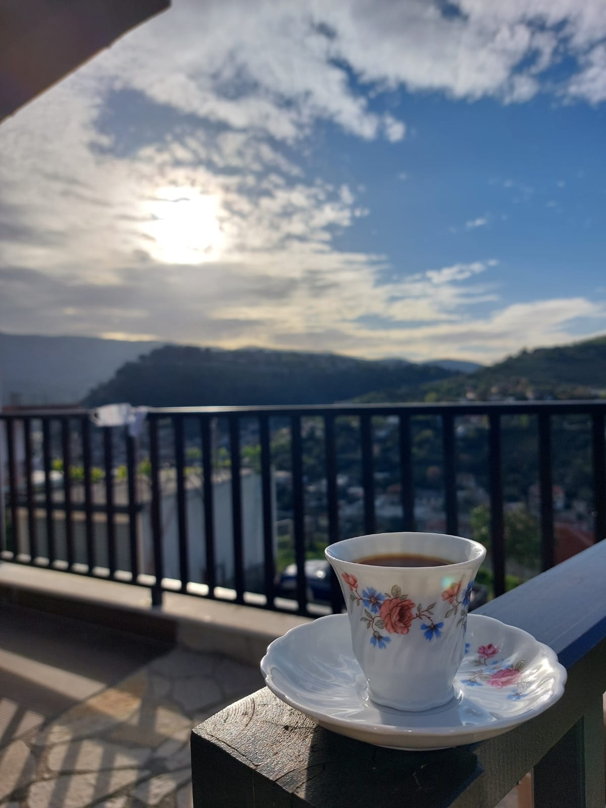 A delicate floral patterned coffee cup rests on a matching saucer, placed on a balcony railing. Expansive views of rolling hills and a cloudy sky are visible in the background, with sunlight filtering through the clouds, creating a serene atmosphere.