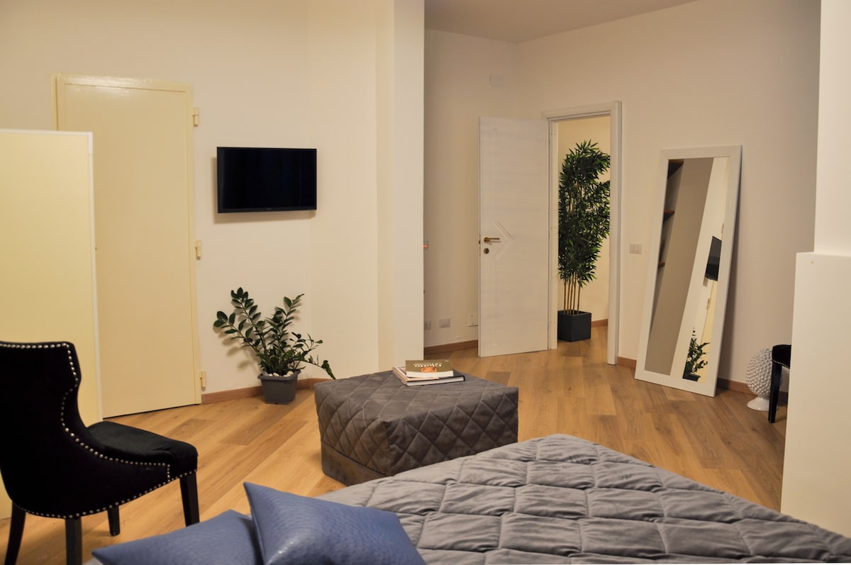 A spacious room is adorned with warm wood flooring. A comfortable bed is positioned in the foreground with soft linens. A large mirror and a lounging chair enhance the decor, while a small potted plant adds a touch of greenery. A wall-mounted television is visible for entertainment.