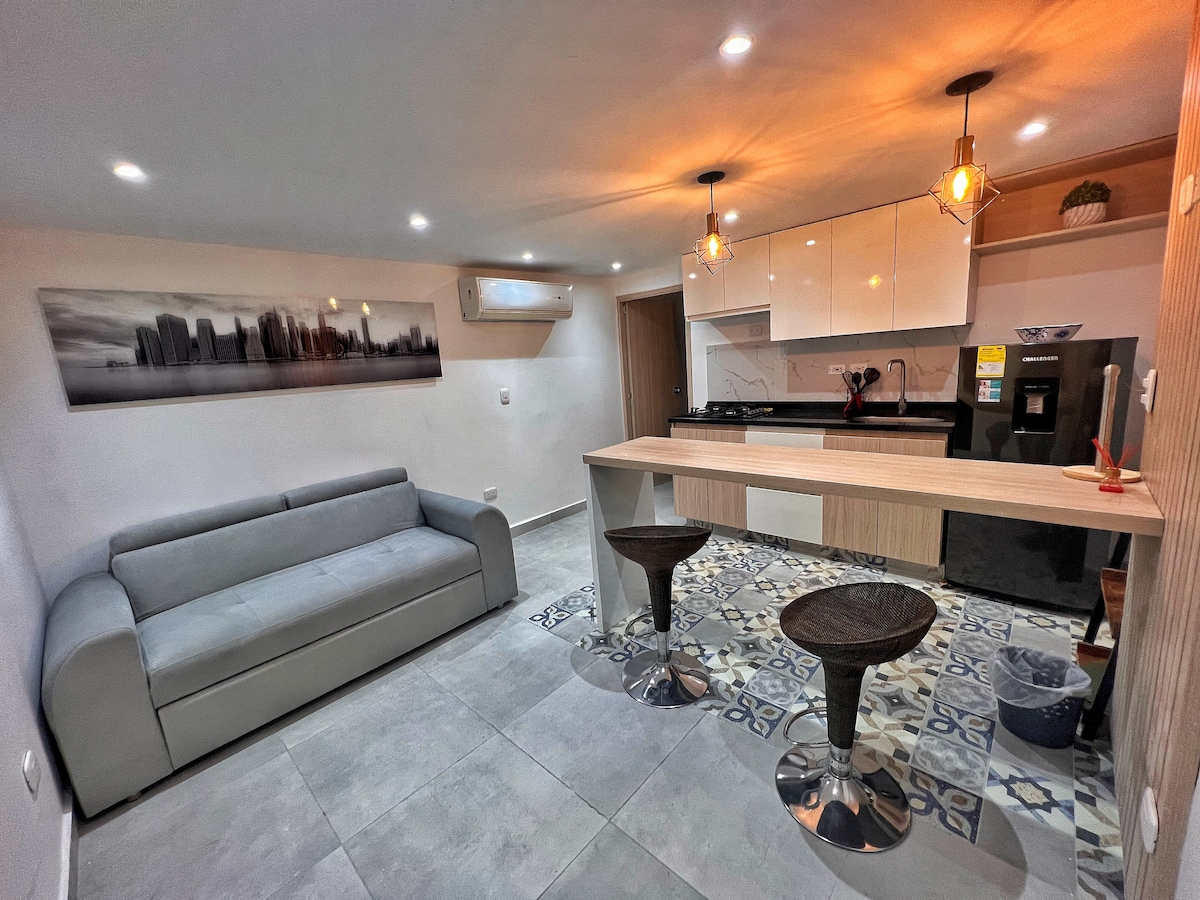 A modern living space features a grey sofa arranged facing a kitchen bar with two stools. The kitchen showcases white cabinetry and contemporary lighting. Decorative tiles on the floor add texture, while wall-mounted artwork complements the ambiance.