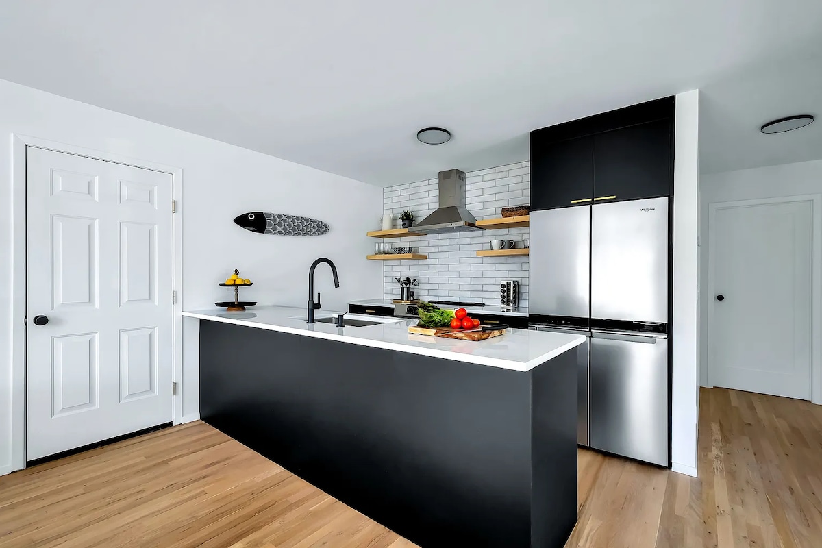 A modern kitchen is presented with sleek black cabinetry and stainless steel appliances. A large island serves as the focal point, topped with fresh fruits and a cutting board. Light wood flooring complements the space, while white walls enhance its brightness.