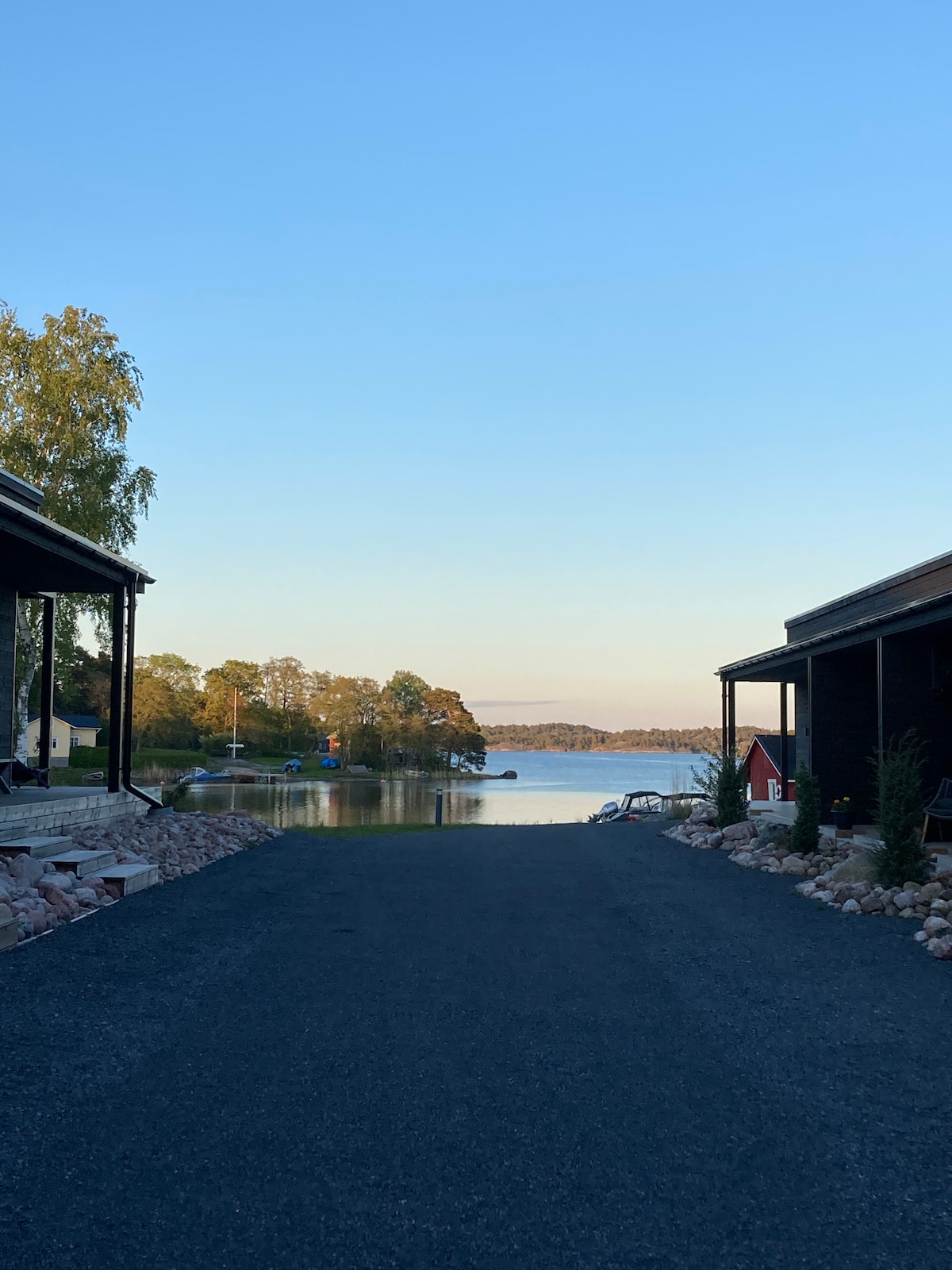 A serene view along a gravel pathway leads to calm waters, framed by wooden structures on either side. The landscape features trees in the background and a glimpse of boats moored nearby, creating a peaceful coastal setting.