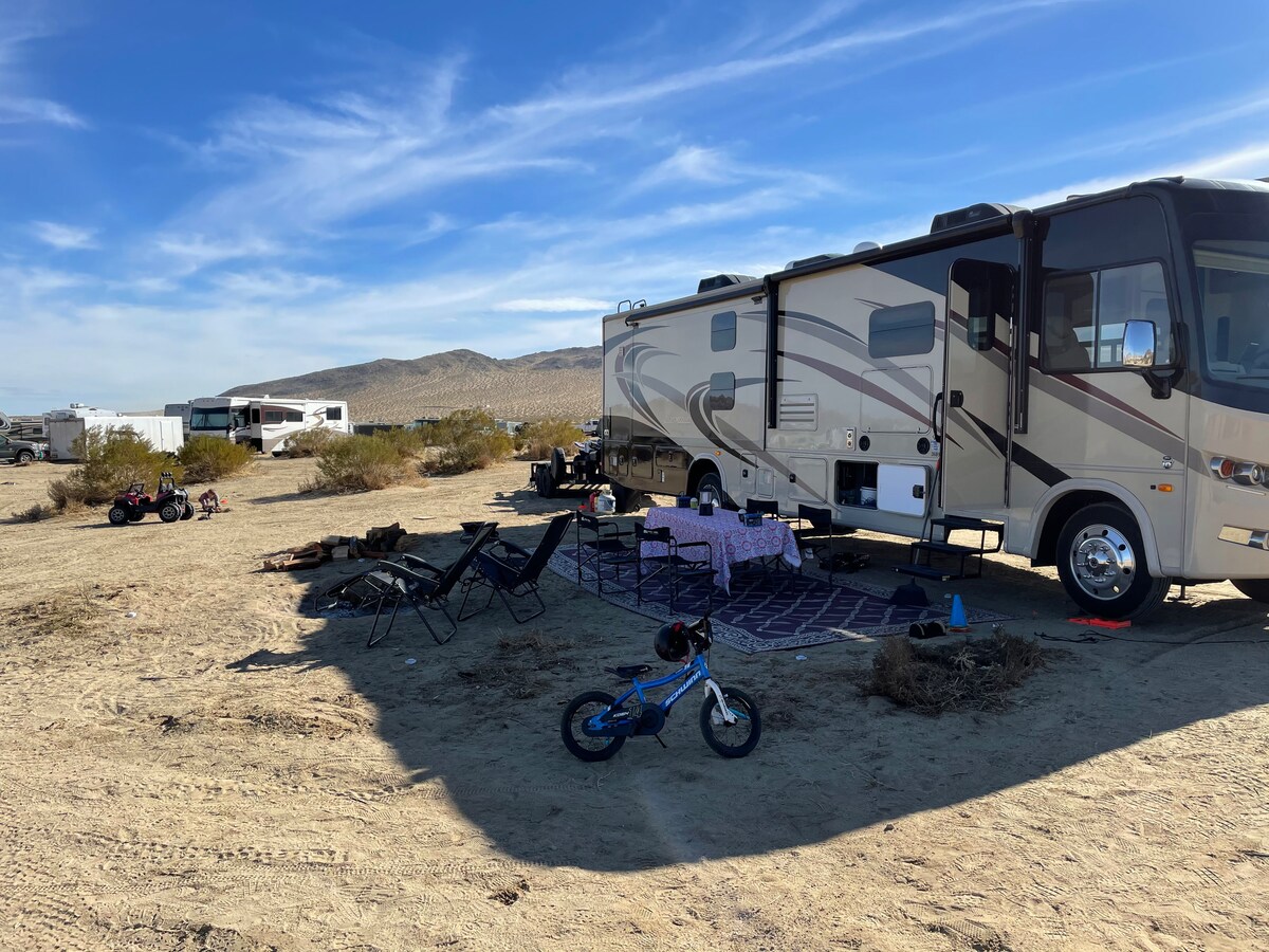 A motorhome is parked on a sandy area, accompanied by outdoor seating, including chairs and a table covered with a patterned tablecloth. A small bicycle is positioned nearby, and distant RVs are visible against a backdrop of desert hills and a clear blue sky.
