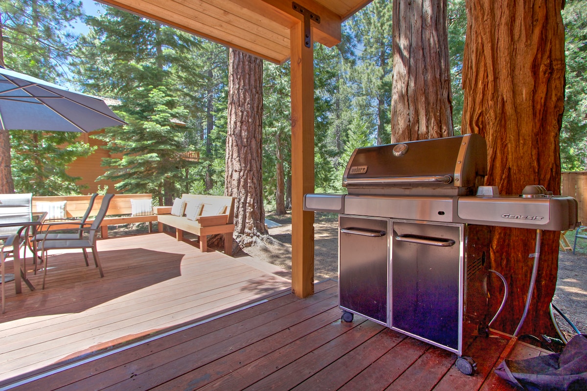 A propane barbecue grill is positioned on a wooden deck, surrounded by tall trees for a natural setting. Patio furniture, featuring a table and chairs, is visible in the background under an umbrella, ideal for outdoor dining.