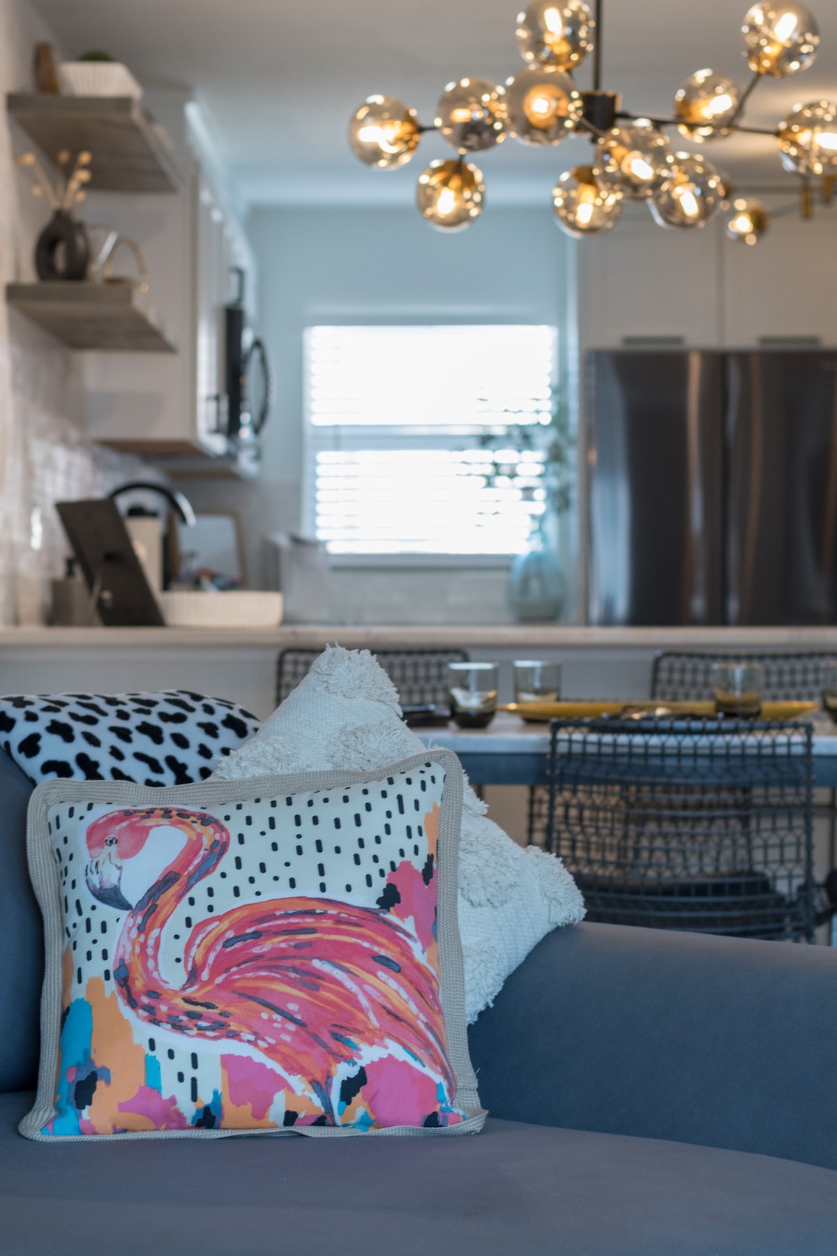 A cozy living area is highlighted by a colorful pillow featuring a flamingo design, resting on a smooth gray couch. In the background, a dining area is visible with a dark table set for four, accompanied by modern chairs. Soft light streams through a nearby window.