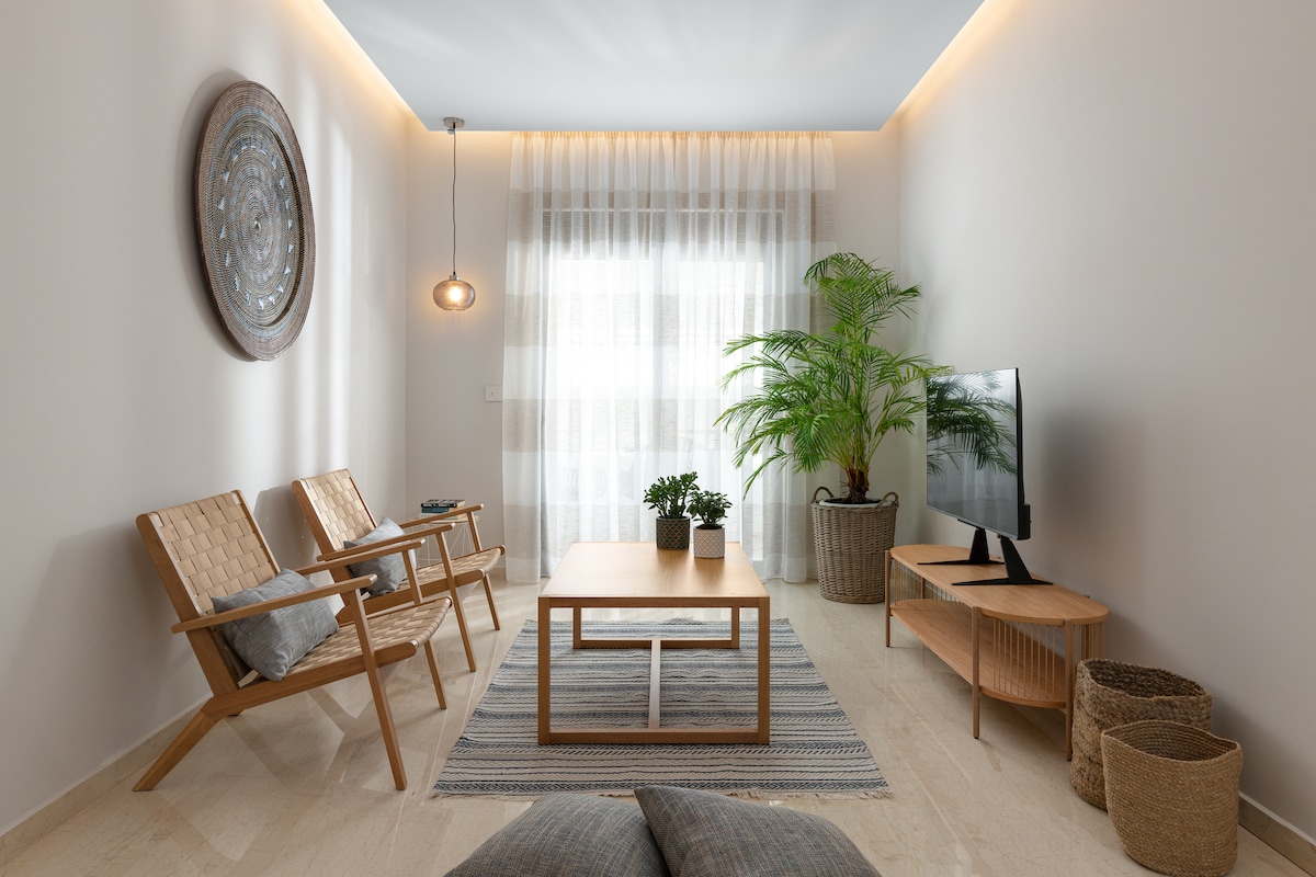 A spacious living area is highlighted by two wooden chairs, a central table, and a television mounted on a wooden console. Natural light filters through sheer curtains, illuminating contrasting plant life in the corner. A patterned rug anchors the space, enhancing its tranquil ambiance.