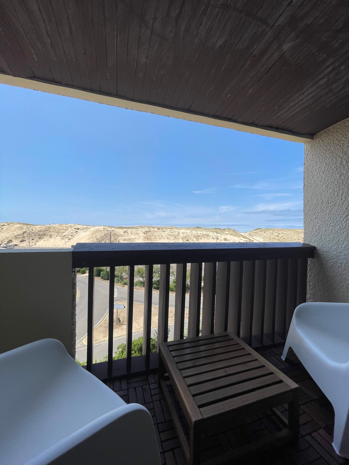 A balcony space is visible, featuring two white chairs and a small wooden table. The outdoor area offers a clear view of sand dunes under a blue sky, creating an open and airy atmosphere.