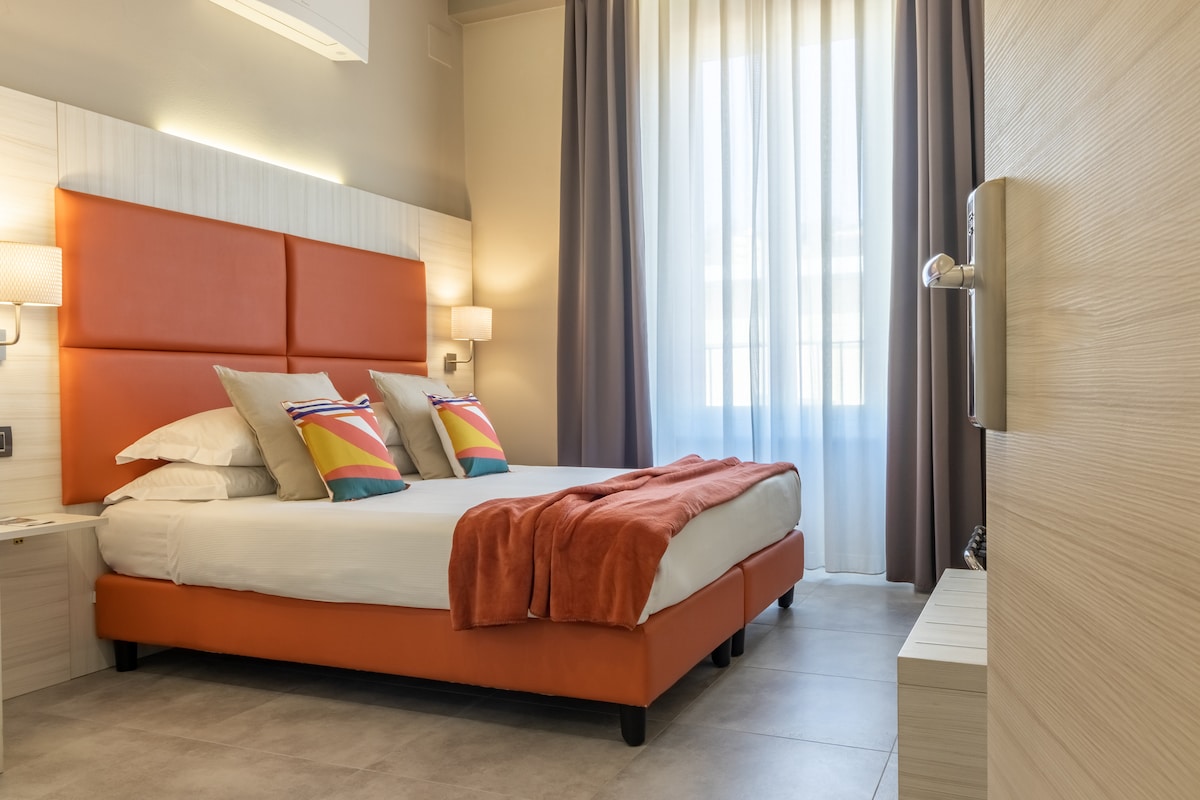 A private room features a cozy bed with vibrant orange headboard and colorful pillows. Soft natural light filters through large curtains, illuminating the minimalist decor and inviting atmosphere. Nightstands with lamps are positioned on either side of the bed, enhancing the room's comfort.