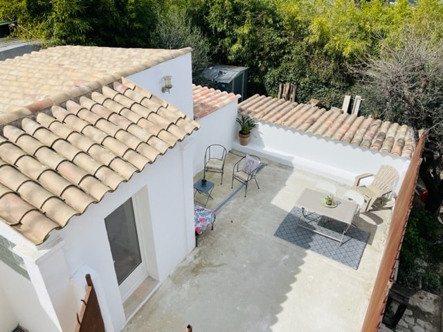 An aerial view of a private terrace featuring a small table and two chairs set against a backdrop of lush greenery. The tiled roof and white walls provide a bright and open feel, while the space appears ideal for relaxation.