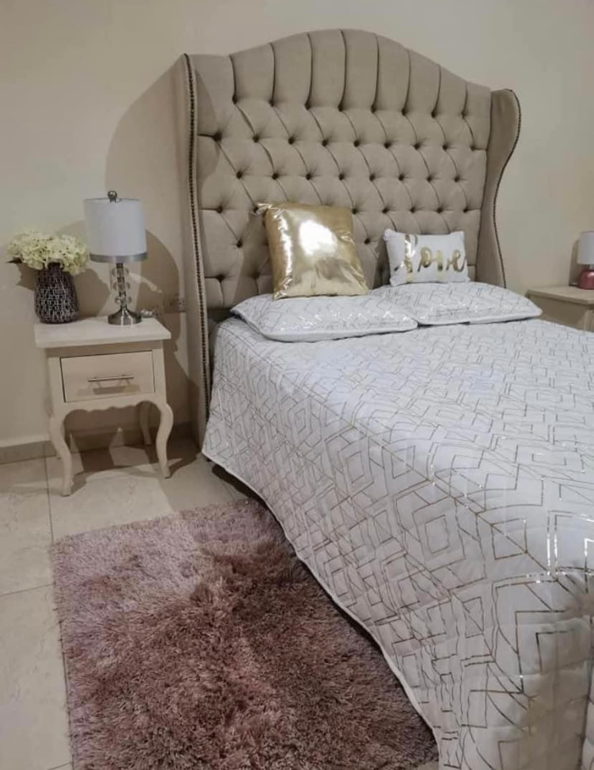 A spacious bed with a tufted headboard is centered in the room, flanked by two nightstands with lamps. Decorative pillows add texture, while a soft rug lies beneath the bed, contributing to a cozy atmosphere.