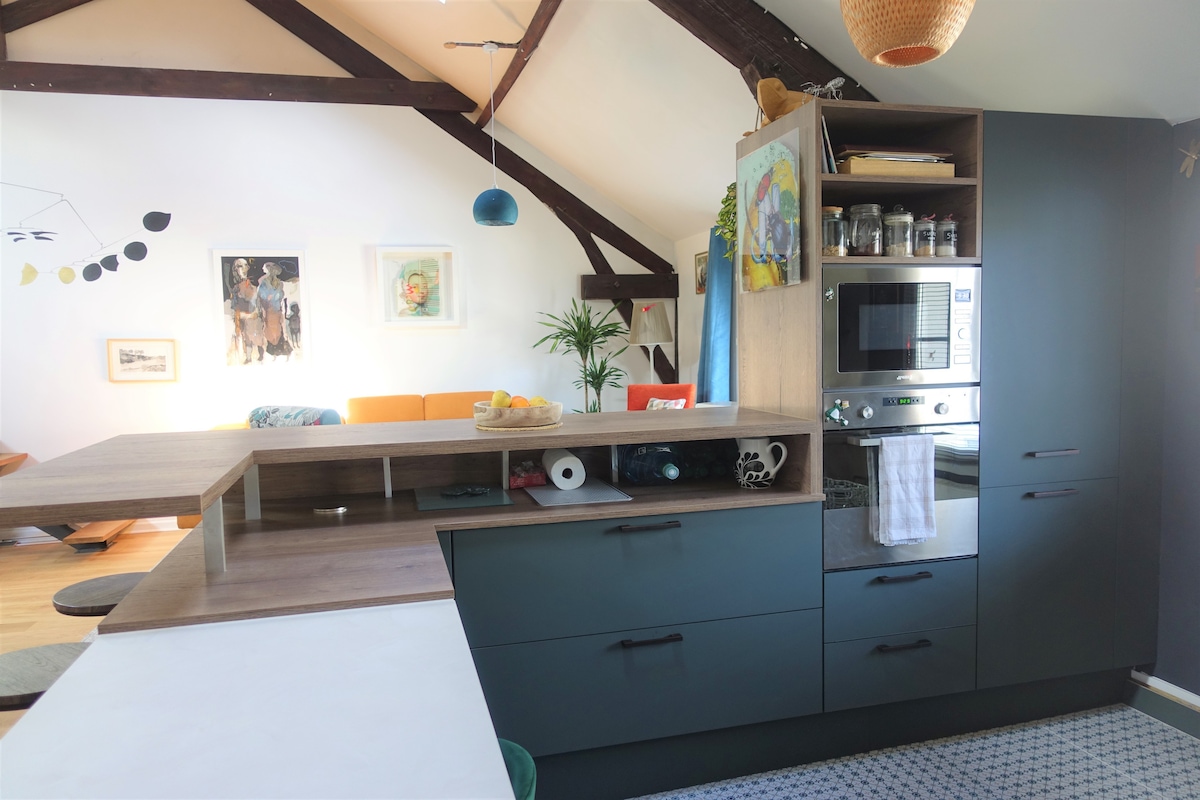 A modern kitchen area features sleek cabinetry in dark tones, with a countertop separating it from the living space. An oven and jars on display add functionality. The high ceilings with exposed beams provide an airy feel, complementing the adjoining sitting area visible in the background.