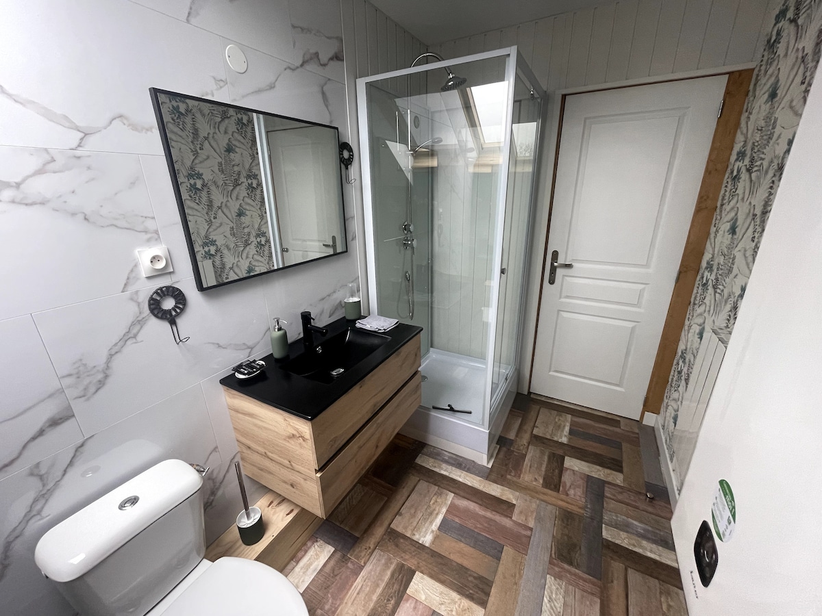 A modern bathroom features a walk-in shower and a sleek wooden vanity with a black sink. A large mirror complements the stylish decor. Natural light illuminates the space, enhanced by patterned wallpaper in a corner. The floor showcases a unique wood design.