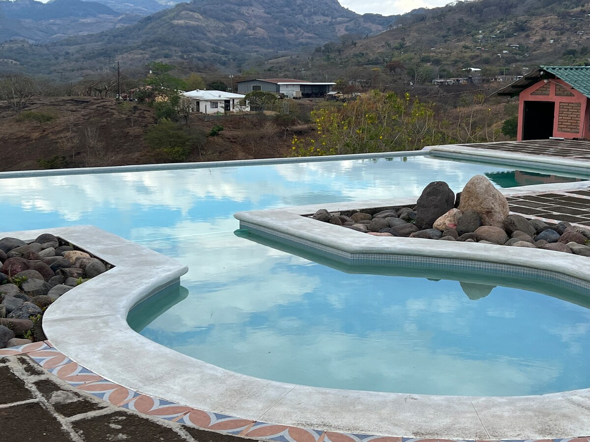 An expansive infinity pool is featured, with calm water reflecting the surrounding landscape. Smooth, light-colored stone edges frame the pool, while natural rocks are positioned artistically nearby. The backdrop consists of rolling hills and a peaceful sky, enhancing the serene ambiance.