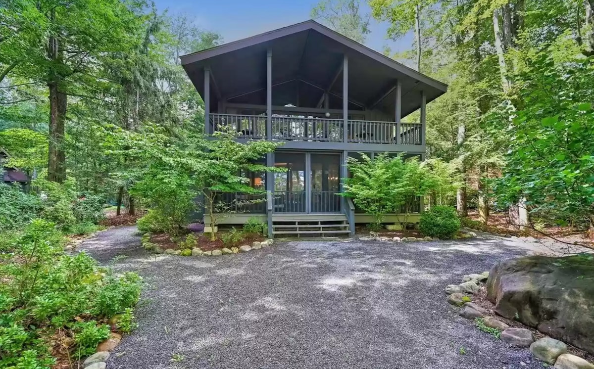 The cabin features a modern design with a spacious porch supported by tall beams, surrounded by lush greenery. A gravel pathway leads to the entrance, which is framed by carefully arranged rocks and vibrant plants, creating a serene outdoor setting.