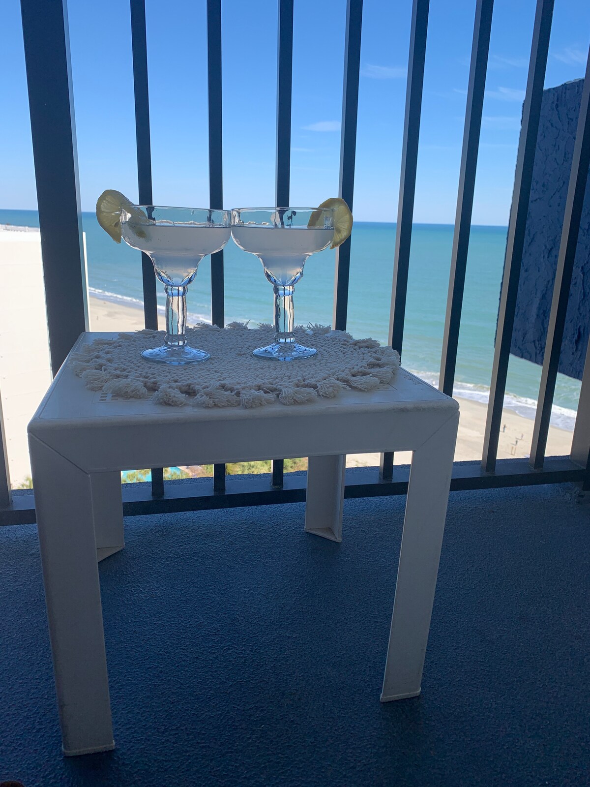 Two glasses of margaritas sit on a small, white table adorned with a decorative mat. A panoramic view of the ocean is visible through the balcony railing, with waves gently lapping the shore under a clear blue sky.