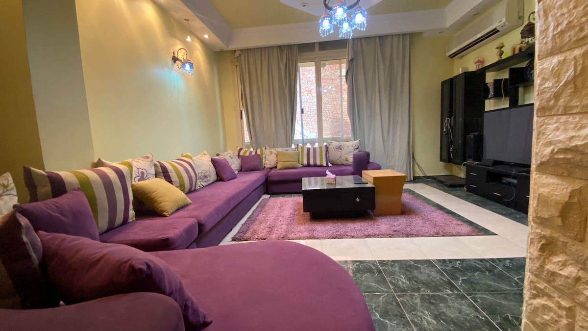 A spacious living room features a large purple sectional sofa with decorative cushions, complemented by a central coffee table and a plush area rug. Soft lighting is provided by elegant ceiling fixtures, while natural light enters through large windows dressed with curtains.