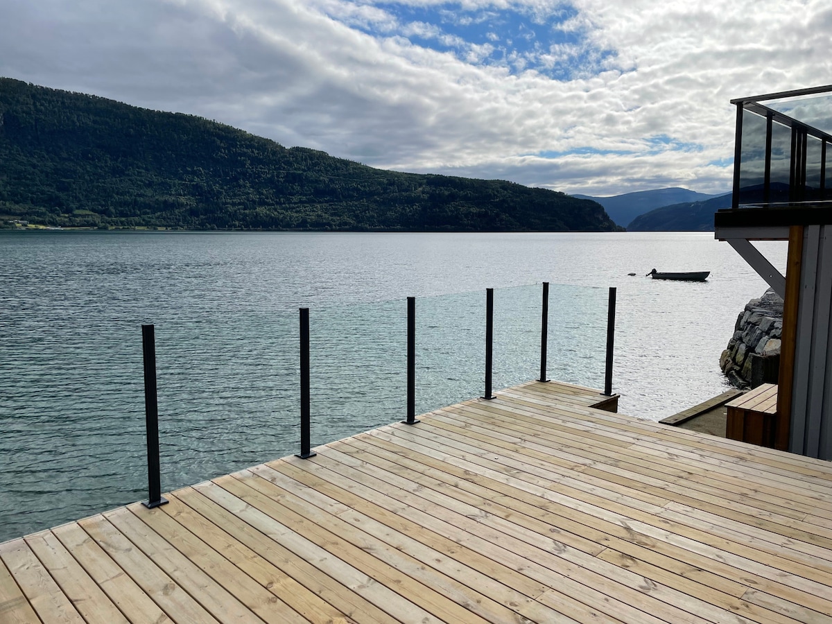A spacious wooden deck extends toward the tranquil waters of the fjord, surrounded by mountains. Glass railings provide unobstructed views of the serene landscape, enhancing the connection to nature. The gentle ripples of the water can be seen reflecting the soft clouds above.