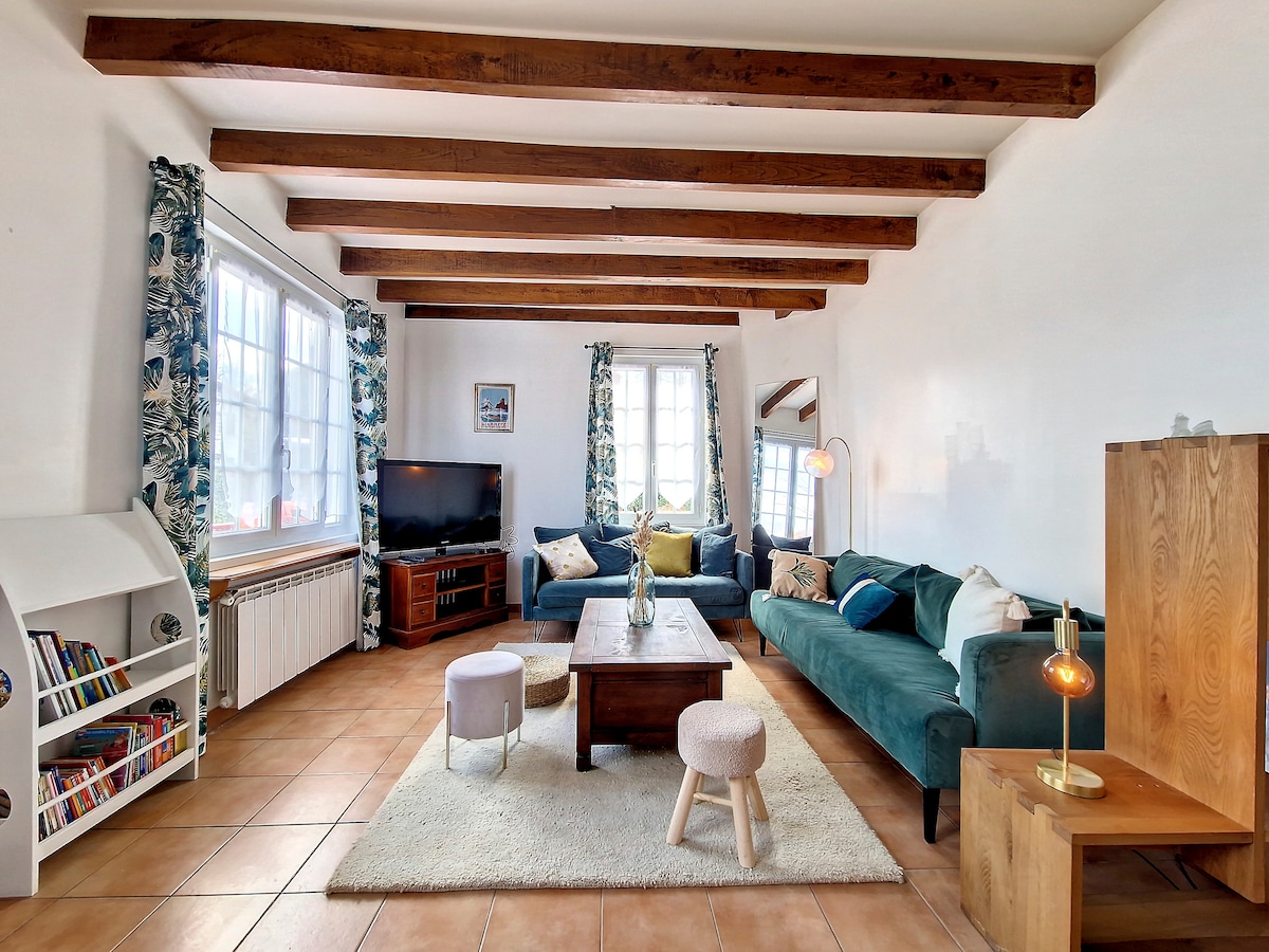 A spacious living area features wooden beams overhead, with a comfortable blue sofa and a cozy armchair. A wooden coffee table is centered on a soft rug, surrounded by light-colored poufs. Large windows allow natural light, and a television is set on a media unit.