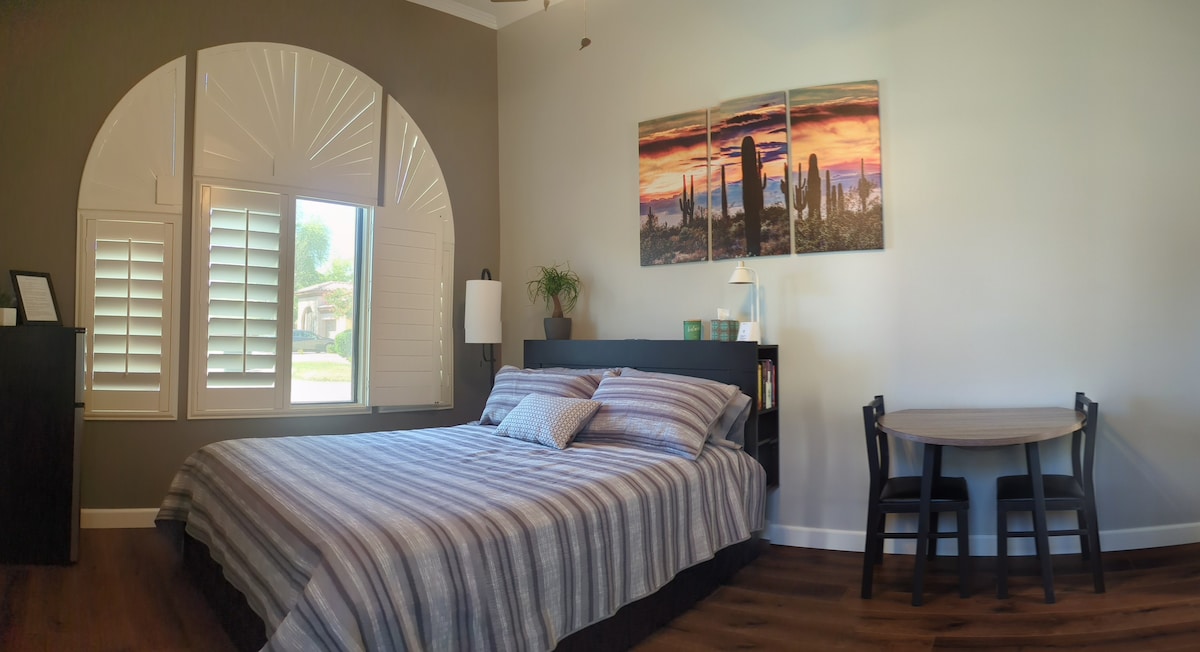 A private room features a spacious bed with layered bedding, complemented by two decorative pillows. A small dining table with two chairs sits in the corner. Large windows with wooden shutters frame a view of greenery outside, and landscape artwork adorns the wall.