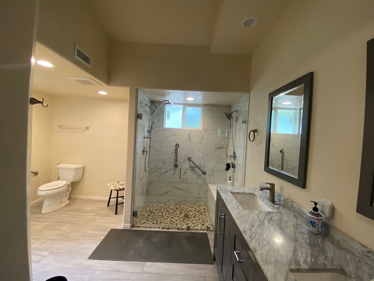 The bathroom features a spacious walk-in shower with multiple showerheads and a pebble stone floor. Natural light enters through two windows, illuminating the light-colored marble walls. A modern vanity with a sink is positioned beside the shower, and a toilet is situated at the far end.