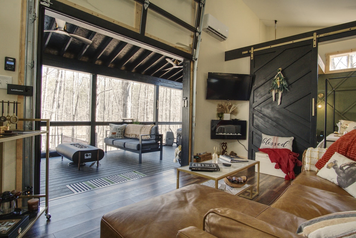 The interior living space features a large leather sofa and a coffee table, with a wall-mounted TV for entertainment. A garage door opens to the screened porch, where comfortable seating and forest views are visible, enhancing the connection to nature.