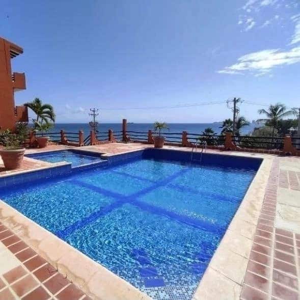 A clear blue pool is set against a backdrop of palm trees and the ocean, reflecting the sky's brightness. Sunlight glistens on the water's surface, while a small area with steps leads into the pool, creating a serene outdoor space for relaxation.