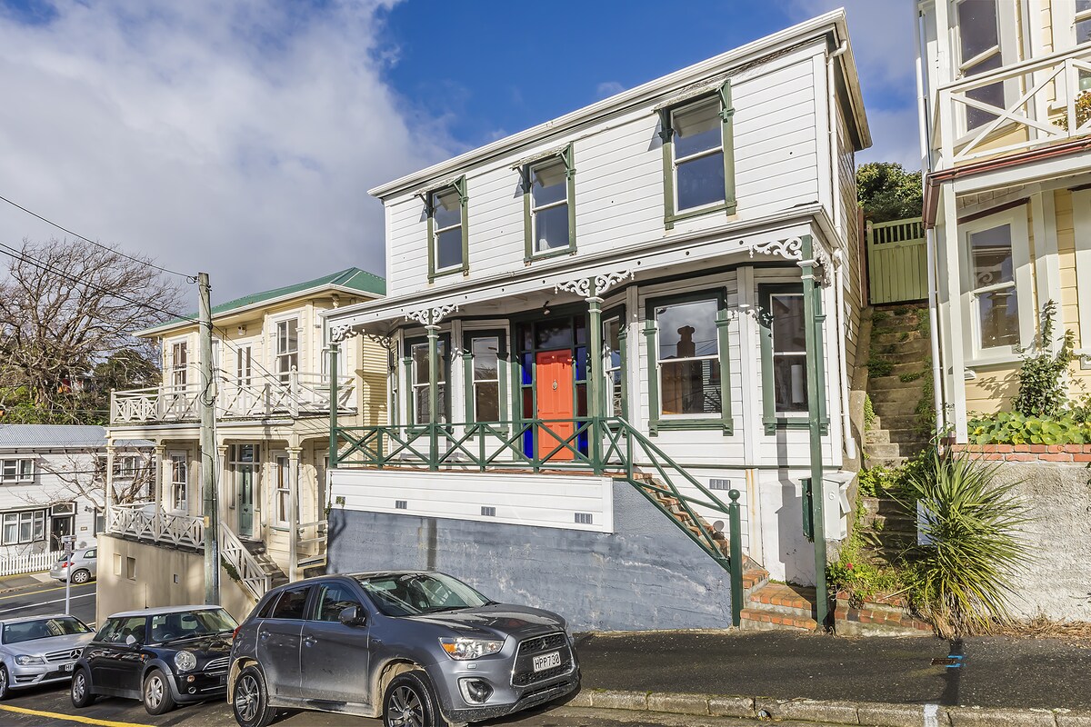 A charming two-story home is presented, featuring a welcoming red front door and decorative trim. The prominent balcony is supported by green railings, and the exterior walls are a light color, contributing to the overall inviting appearance. Several parked vehicles are visible in the foreground.