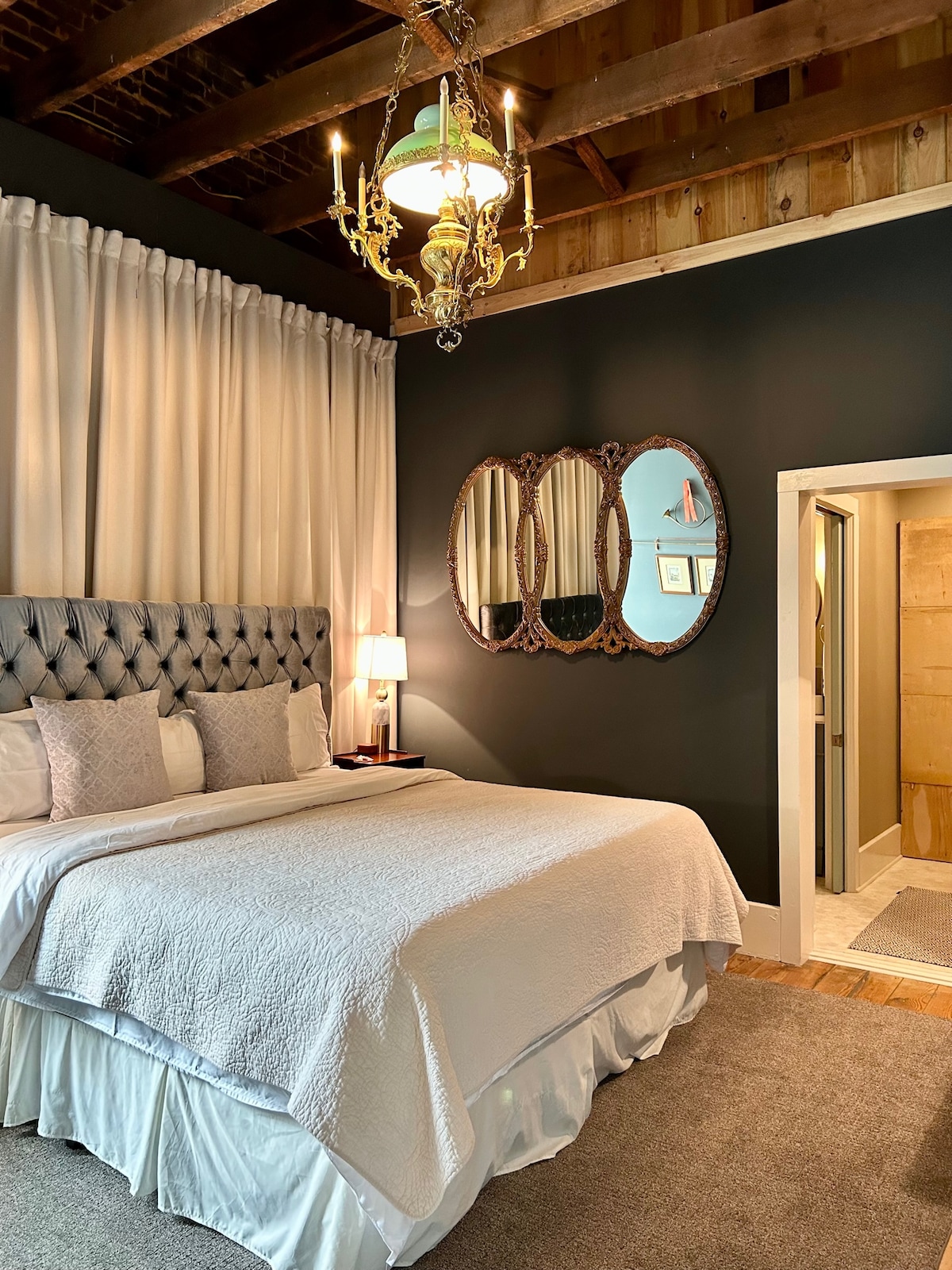 A comfortable bedroom features a spacious bed with a light quilt and decorative pillows. Elegant mirrors are mounted on a dark wall, and a chandelier hangs above. Light streams in through sheer curtains, illuminating the cozy space with a warm glow. A doorway leads to the private bathroom.
