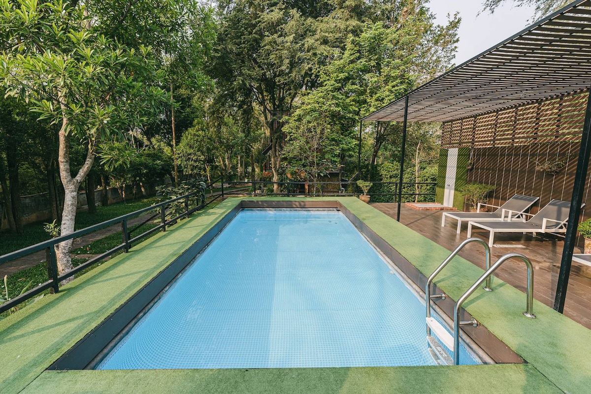 A serene swimming pool is surrounded by lush greenery, with a wooden deck offering seating options nearby. The clear blue water reflects the natural surroundings, enhancing the tranquil atmosphere of the space.