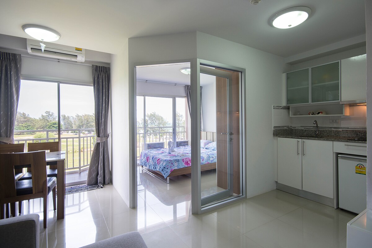 An open-plan living space features a contemporary kitchen with sleek cabinetry. A dining table for four is positioned near large windows, providing views of the outdoors. A cozy sleeping area is separated by glass doors, with a double bed adorned in floral bedding.